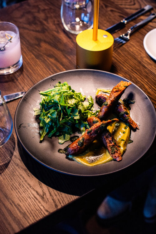 Grilled sausages with puree and greens on a black plate, served with a pink cocktail.