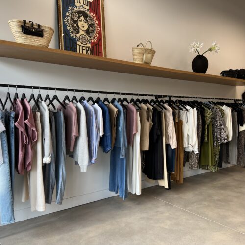 Clothing rack in a boutique with neatly arranged casual and stylish outfits, under a shelf with bags, flowers, and artwork.